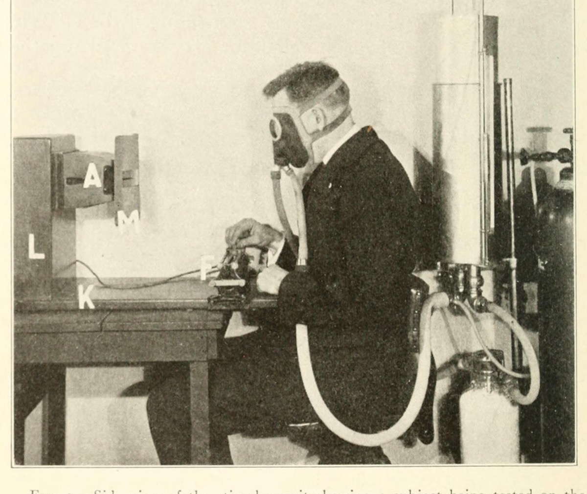 Black-and-white laboratory photograph of a man in a breathing mask seated at a table with an electrical meter.