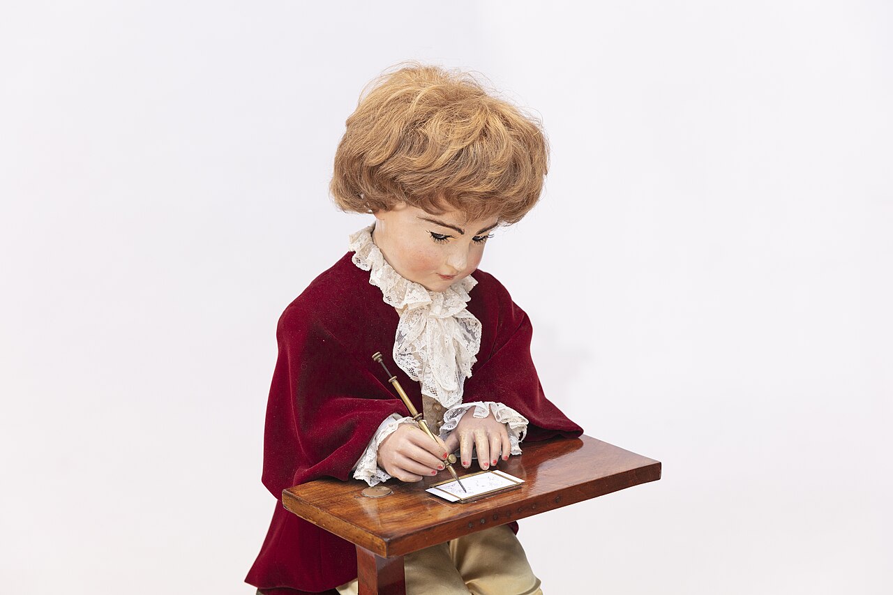 A clockwork automaton of a curly-haired boy in a red velvet coat, bent over a small desk, drawing with a pencil.