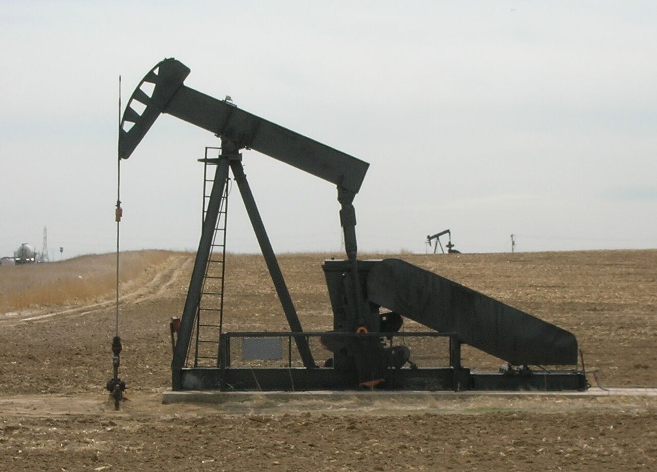A black oil-well pump jack standing in a brown field under a pale sky.